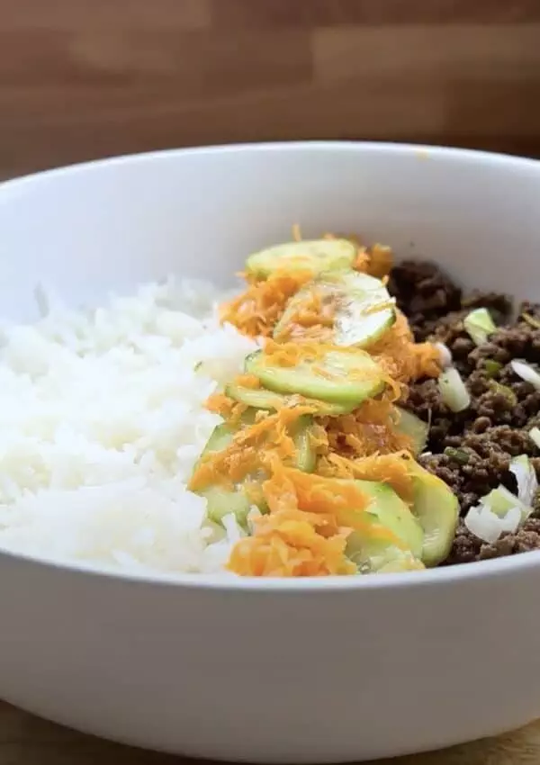 Rice bowl with ground beef, sliced cucumbers, and grated carrots in a white bowl on a wooden table.